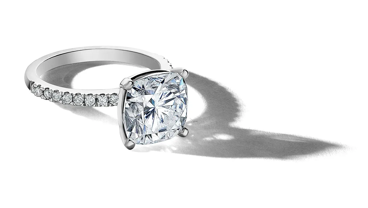 A stunning engagement ring designed by Ex Aurum Jewelers, featured in the wedding jewelry category on Mariage Elegant, Canada. The ring showcases a large, brilliant cushion-cut diamond set in a classic four-prong setting. The band is adorned with a series of smaller round diamonds, enhancing its elegance and sparkle. The image captures the ring on a reflective surface, highlighting its exquisite craftsmanship and luxurious appeal, perfect for couples planning their special day.