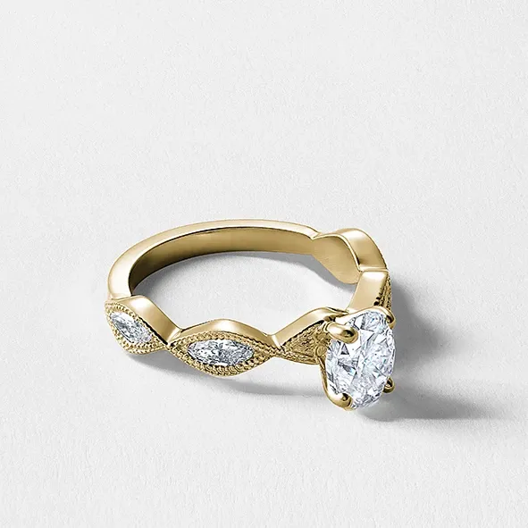 The image features an elegant engagement ring from Ex Aurum Jewelers, showcased under the wedding jewelry category. The ring is crafted from polished gold with a unique band design that includes marquise-shaped elements accented with sparkling diamonds. At its center is a prominent oval-cut diamond, exuding sophistication and brilliance, perfect for a wedding proposal. The ring rests against a stark white background, highlighting its intricate details and luxurious finish, ideal for couples seeking exquisite jewelry for their special day.