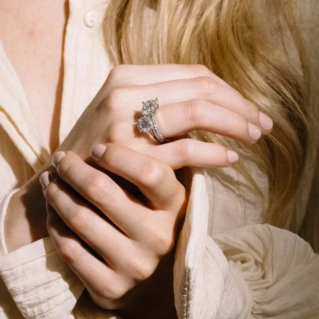 A woman's hands are gracefully clasped against a soft fabric backdrop, showcasing an intricately designed engagement ring and wedding band from Ex Aurum Jewelers. The engagement ring features a prominent, sparkling diamond at its center, complemented by smaller diamonds along the band. The matching wedding band enhances the elegance of the set, both perfect for a sophisticated wedding jewelry choice.