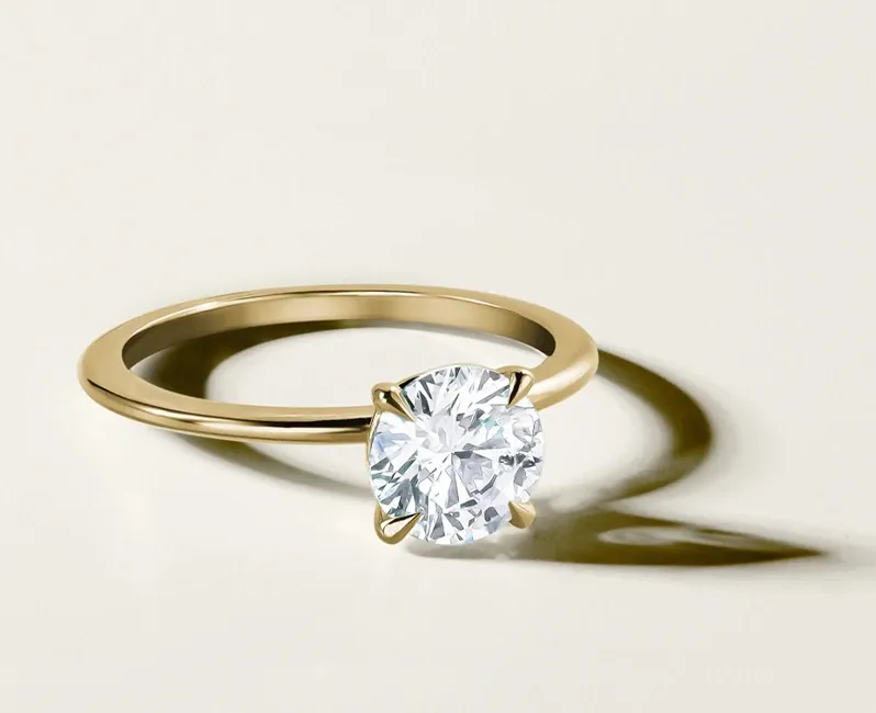 A close-up image of an elegant engagement ring by Ex Aurum Jewelers, featured in the Wedding Jewelry category of Mariage Elegant, Canada. The ring displays a brilliant, round-cut diamond set in a classic yellow gold band. The diamond's facets are precisely cut, enhancing its sparkle and capturing light beautifully. The simple yet sophisticated design of this piece makes it a perfect choice for those planning their wedding and seeking timeless jewelry. The warm tones of the gold band complement the clarity of the diamond, offering a luxurious and refined aesthetic ideal for a wedding celebration.