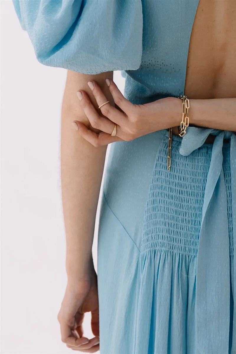 A bride wearing a light blue dress showcases elegant gold jewelry from Ex Aurum Jewelers, a vendor specializing in wedding jewelry. The image highlights a delicate gold bracelet and a simple yet elegant gold ring on her fingers. The dress features a smocked waist and puffed sleeves, adding a touch of romance. This visual captures the essence of refined wedding accessories for Mariage Elegance, Canada.