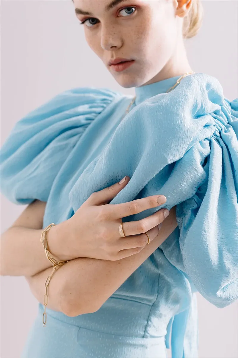 A woman wearing an elegant blue dress with voluminous sleeves is showcasing jewelry from Ex Aurum Jewelers, featured in the Wedding Jewelry category on Mariage Elegant. She adorns a delicate gold bracelet and a simple gold ring, adding a touch of sophistication to her outfit. The image highlights the intricate design and craftsmanship of the jewelry, ideal for wedding celebrations and special occasions.