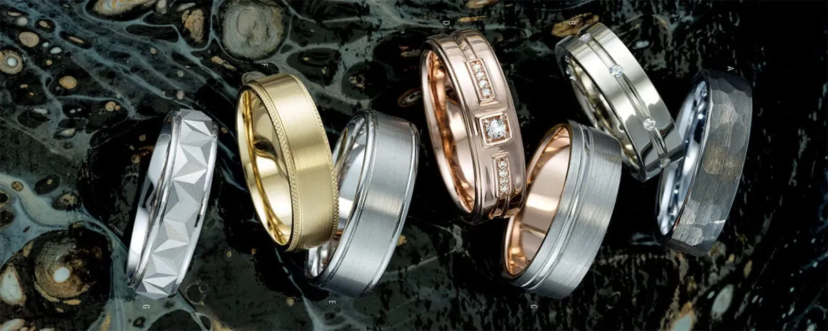A diverse array of wedding rings from Ex Aurum Jewelers, featured under the Wedding Jewelry category on Mariage Elegant, Canada. The image showcases seven exquisite rings arranged in a visually pleasing manner against a marbled, dark surface. Each ring displays unique craftsmanship: a faceted geometric design, a smooth polished gold band, a brushed silver band, a detailed band with square diamond settings, a classic silver band with subtle grooves, a textured silver ring with a hammered finish, and a sleek silver ring with an intricate pattern. These rings reflect the elegance and sophistication perfect for wedding celebrations.