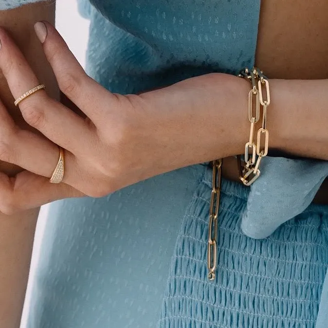 A close-up image of a woman's hands adorned with elegant wedding jewelry from Ex Aurum Jewelers, categorized under Wedding Jewelry for Mariage Elegant in Canada. She is wearing a delicate gold chain bracelet with a modern geometric design and coordinating gold rings that enhance the refined sophistication of her attire. The light blue textured fabric of her dress adds a soft contrast, emphasizing the intricate details of the jewelry pieces, ideal for brides seeking timeless elegance.