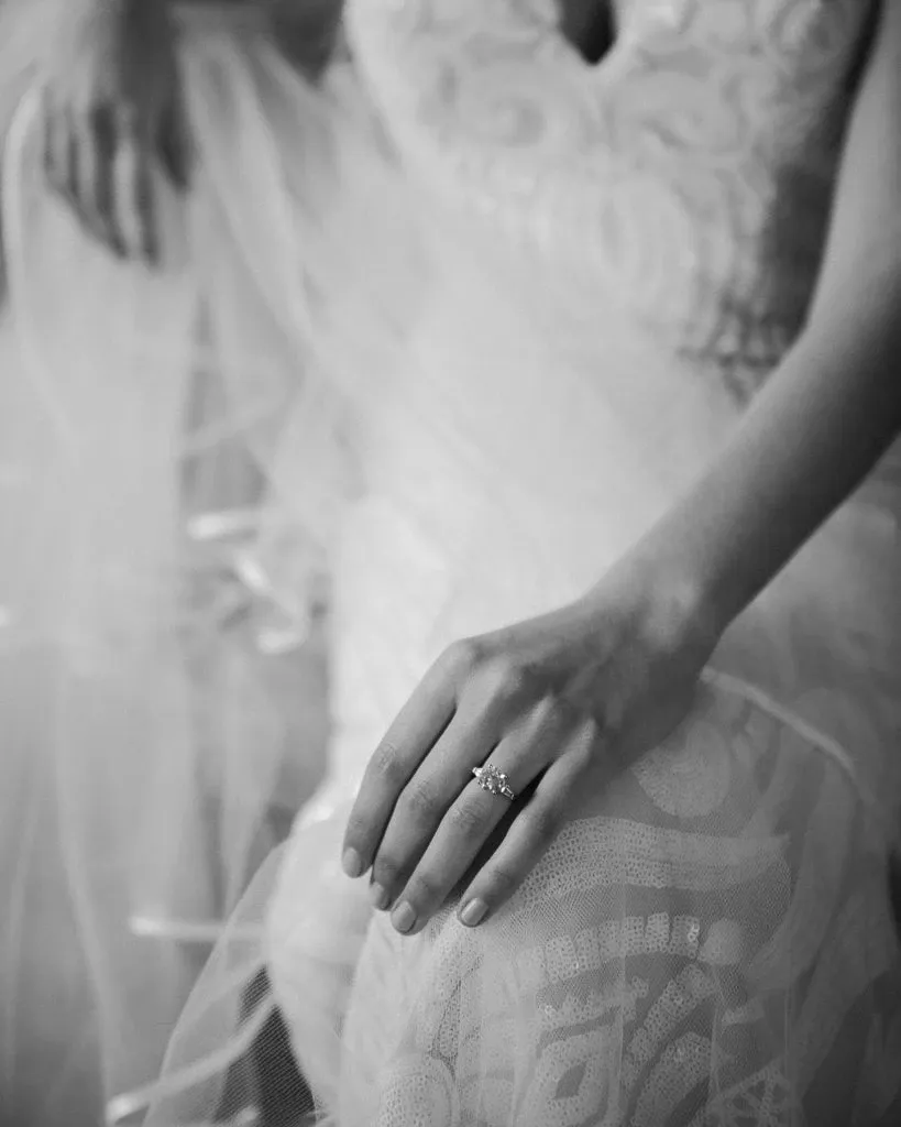 This black and white image features a bride wearing a delicate lace wedding dress, seated gracefully with her hand resting on her lap. The focus is on a stunning diamond engagement ring from Ex Aurum Jewelers, a vendor in the Wedding Jewelry category. The intricacy of the lace detailing on the dress and the elegant design of the ring highlight the sophistication and elegance associated with wedding preparations, as showcased by Mariage Elegant in Canada.