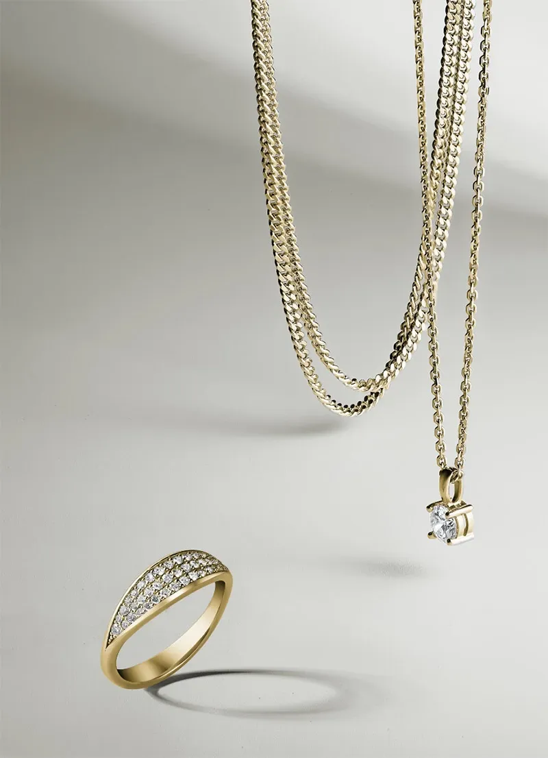 The image showcases a collection of exquisite wedding jewelry by Ex Aurum Jewelers, featured in the Wedding Jewelry category on Mariage Elegant, Canada. The foreground displays an elegant gold ring, beautifully embellished with rows of sparkling diamonds. Hanging alongside the ring are two delicate gold necklaces, one featuring a shimmering solitaire diamond pendant. The jewelry pieces rest on a soft, neutral background, highlighting their luxurious craftsmanship and sophisticated designs, ideal for any wedding ensemble.