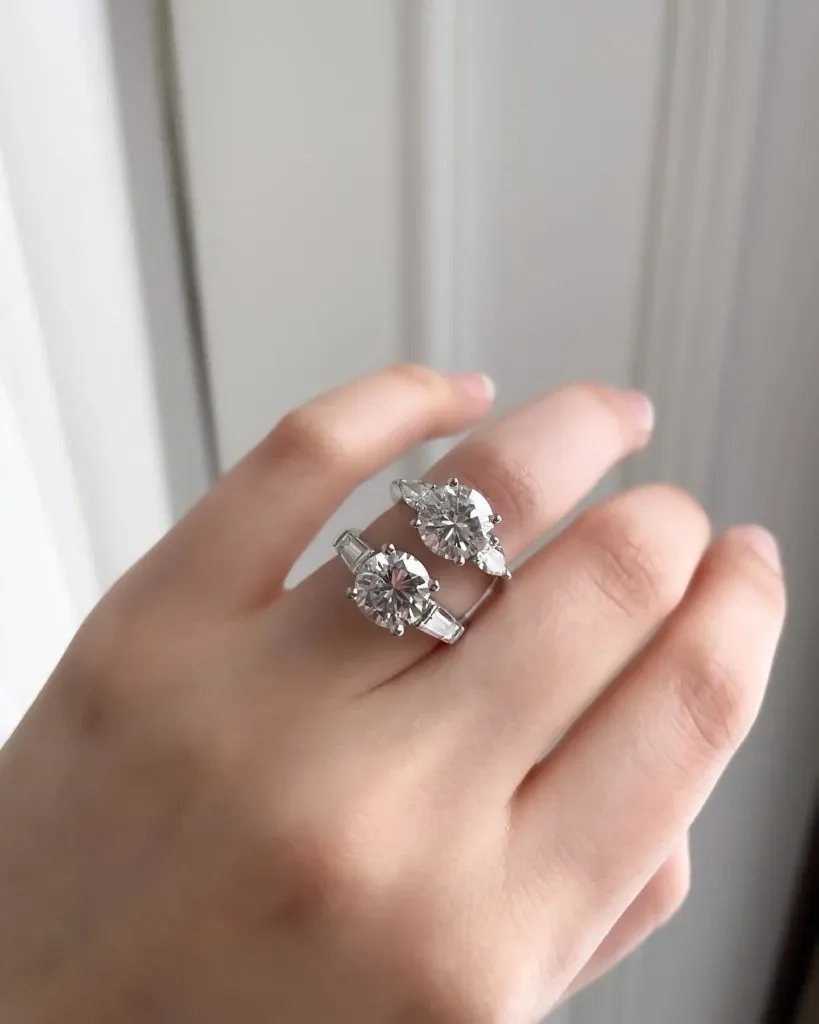 A close-up image showcases two exquisite diamond rings from Ex Aurum Jewelers, fitting perfectly within the Wedding Jewelry category. The rings are elegantly displayed on a person's hand, highlighting their intricate designs and sparkling gemstones. The first ring features a round-cut diamond set in a classic band, while the second ring displays an oval-cut diamond with additional stones set along the band, emphasizing luxury and sophistication for wedding occasions. The backdrop of the image is a soft, neutral-colored surface that enhances the brilliance of the jewelry pieces, making them ideal choices for an elegant wedding ceremony featured on Mariage Elegant, Canada.
