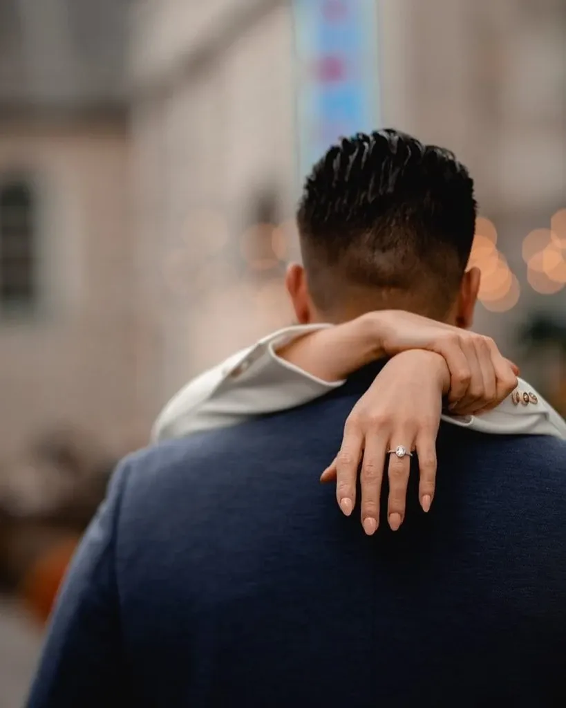 In this image for Mariage Elegant's wedding vendor listing, a couple embraces, showcasing a stunning engagement ring from Ex Aurum Jewelers in the Wedding Jewelry category. The focus is on the bride's left hand, elegantly draped over the groom's shoulder, highlighting the beautiful diamond ring set against a soft blurred background. The groom is dressed in a dark blue suit, and the scene exudes a romantic and intimate atmosphere, perfect for a wedding setting.