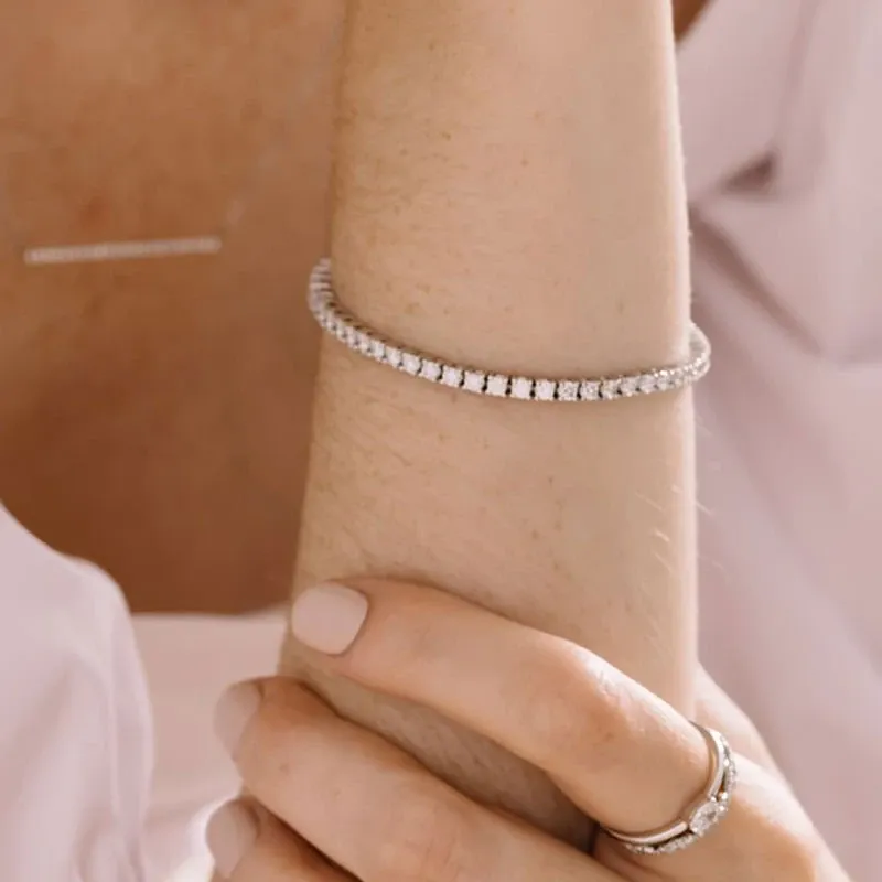 A woman elegantly wearing wedding jewelry by Ex Aurum Jewelers. The image showcases a delicate diamond tennis bracelet adorning her wrist and a matching diamond ring on her finger. Her nails are neatly manicured with a soft, natural polish. In the background, a subtle, linear diamond necklace can be seen, enhancing the sophisticated theme, typical of the wedding jewelry category from Mariage Elegant, Canada.