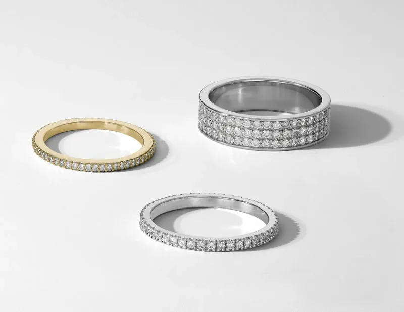 The image features a collection of exquisite wedding jewelry from Ex Aurum Jewelers, showcasing three elegant rings. On the top right is a silver band encrusted with multiple rows of sparkling diamonds, exuding luxury and sophistication. In the foreground, two slender rings are displayed; one is a simple yet elegant band crafted from gold with a single row of diamonds, while the other mirrors the sophisticated style of the first, featuring a silver band with a single row of dazzling stones. These rings are perfect for couples seeking timeless elegance for their wedding day.