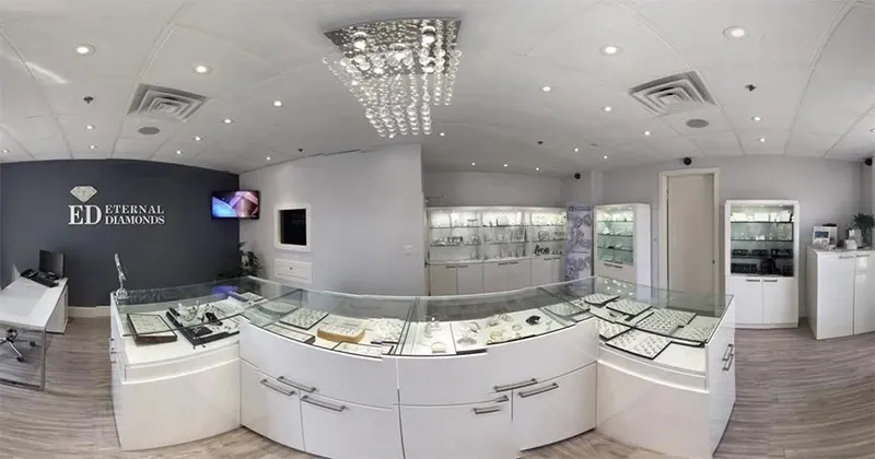 The image shows the interior of Eternal Diamonds, a vendor in the wedding jewelry category. The elegant showroom features a modern design with sleek white display cases showcasing a wide array of sparkling jewelry pieces, including rings and necklaces, under soft lighting. In the center, a curved glass display table highlights more exquisite items, creating a luxurious atmosphere ideal for wedding preparations. The wall behind the displays prominently features the Eternal Diamonds logo, reinforcing the brand's presence. A crystal chandelier hangs from the ceiling, adding a touch of sophistication.