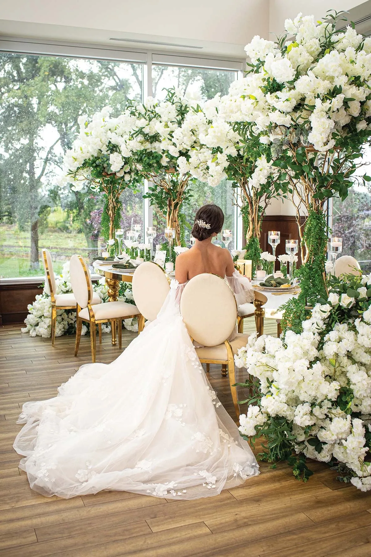 In this elegant wedding decor image by Casa D’Eramo, a bride is seated at a luxuriously decorated table surrounded by lush floral arrangements. The scene features tall, cascading white blossoms with verdant greenery elegantly entwined around golden branches, creating a romantic and opulent atmosphere. The bride's long, flowing gown is beautifully draped over the wooden floor, accentuating the sophisticated setting inside a light-filled room with large windows offering views of the serene greenery outside. Candles and crystal glassware on the table add a touch of elegance, highlighting Casa D’Eramo's exquisite taste in wedding decor.
