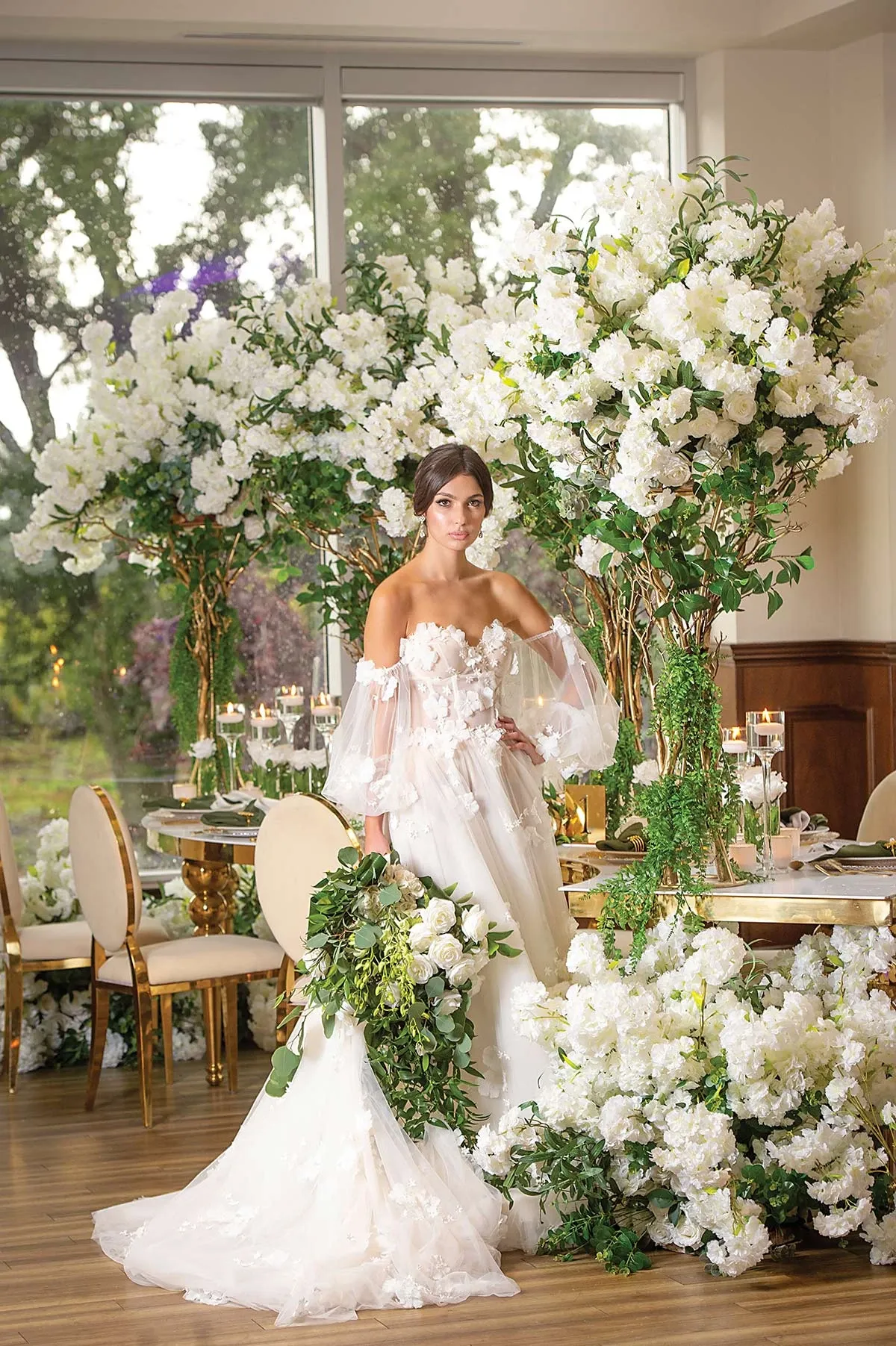 The image showcases an elegant wedding decor setup by Casa D’Eramo in the Wedding Decor category for Mariage Elegant, Canada. The setting features a bride in a beautiful white gown with intricate floral details, surrounded by opulent arrangements of lush white flowers and greenery. The decor includes tall floral towers and cascading flowers on the floor, creating a luxurious and enchanting atmosphere. In the background, large windows offer a view of greenery outside, enhancing the natural and sophisticated ambiance of the wedding venue. The room is adorned with gold-accented dining chairs and softly lit candles on the tables, adding to the intimate and romantic feel.