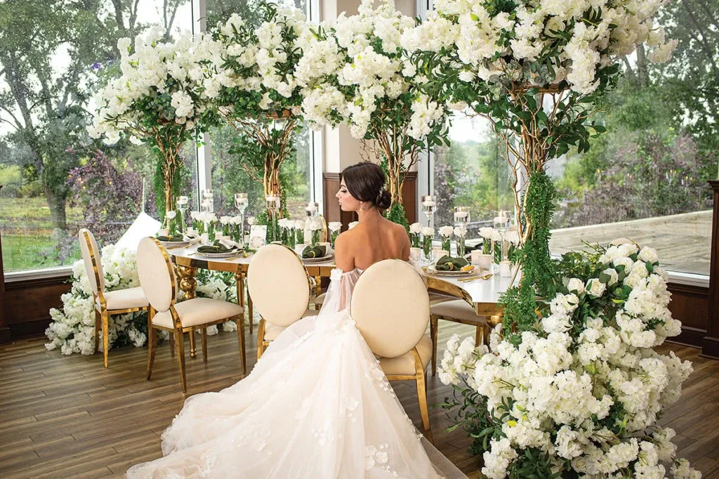 This image features an elegant wedding decor setup by Casa D’Eramo, categorized under Wedding Decor on Mariage Elegant, Canada. A bride in a flowing white gown sits at a finely set table adorned with lush arrangements of white flowers and greenery. The decor includes tall floral installations that frame the table, creating an intimate and luxurious atmosphere. Gold-accented chairs complement the sophisticated theme, and tall candles add a romantic touch. Large windows in the background reveal a serene outdoor view, enhancing the overall setting with natural light.