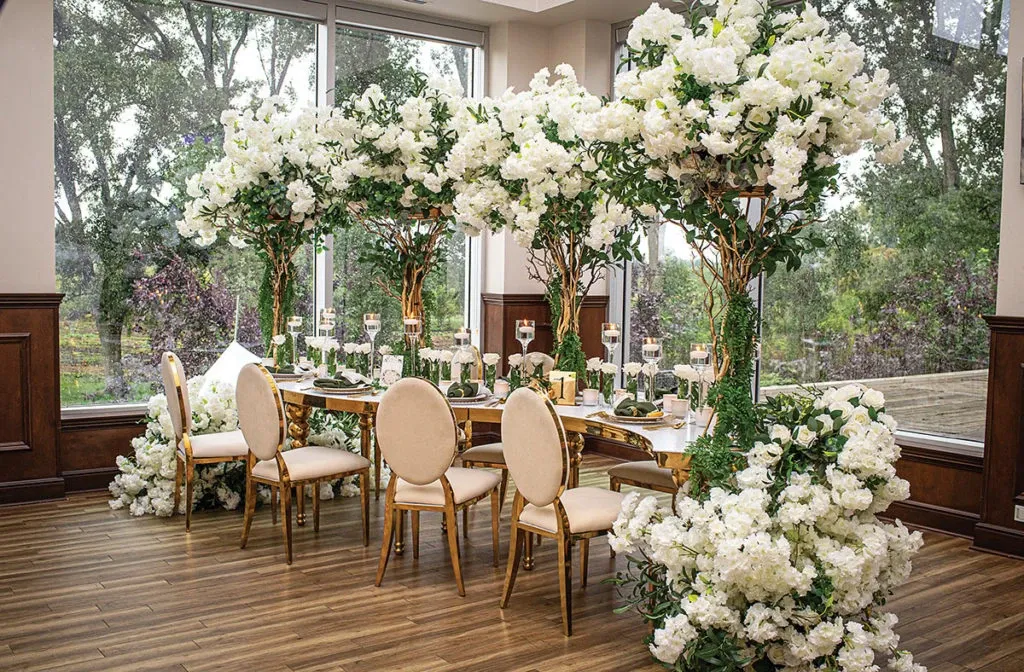 The image features an elegantly decorated wedding reception area by Casa D’Eramo, categorized under Wedding Decor. The setting includes a beautifully arranged dining table surrounded by plush, beige upholstered chairs with gold frames. The table is adorned with intricate centerpieces made of white and green floral arrangements, extending upwards to create a canopy effect. The arrangements consist of lush white flowers and verdant greenery, adding a touch of elegance and sophistication. Tall, elegant candleholders with lit candles are placed on the table, enhancing the romantic ambiance. The venue is framed by large glass windows that provide a view of lush greenery outside, contributing to the serene and picturesque setting. This display showcases Casa D’Eramo’s expertise in creating luxurious and memorable wedding environments.