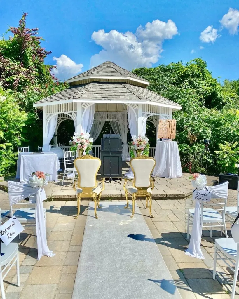 The image showcases an elegant outdoor wedding setup by Casa D’Eramo in the wedding decor category. A beautifully decorated white gazebo serves as the focal point, surrounded by lush greenery and vibrant foliage. Inside the gazebo, there are floral arrangements on white draped tables, adding a touch of romance. In front of the gazebo, two ornate golden chairs are prominently placed facing the aisle. The aisle is lined with white chairs adorned with fabric and floral accents, creating a harmonious and sophisticated atmosphere. The clear blue sky with fluffy clouds enhances the serene and joyful setting, ideal for a wedding ceremony.
