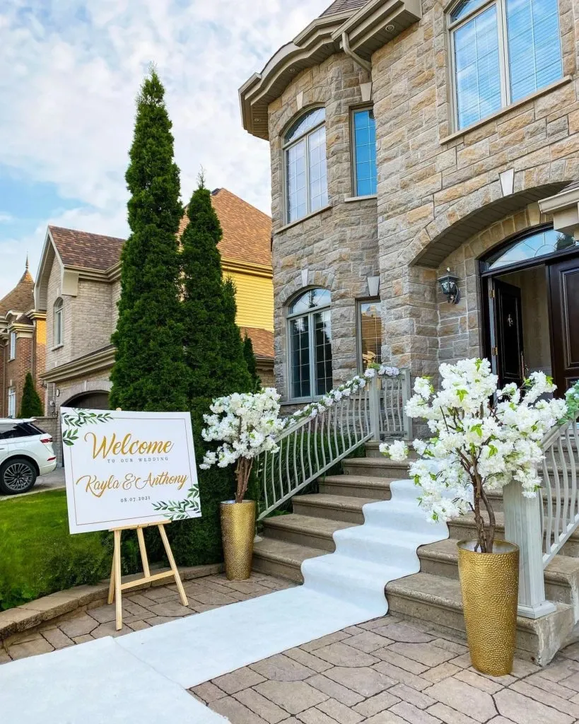 The image shows the elegant exterior of a stone house decorated for a wedding by Casa D'Eramo. In the foreground, there is a white aisle runner leading up the steps to the entrance. On either side of the entrance, tall gold vases hold arrangements of white blossoms, adding a touch of sophistication. A large, welcoming sign on the left reads 'Welcome to the wedding of Kayla & Anthony,' with the date '05.30.2021' inscribed below. This setup by Casa D'Eramo is a beautiful example of their wedding decor expertise, perfect for elegant and memorable ceremonies.