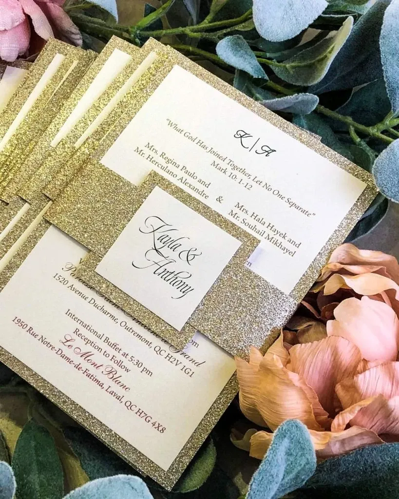 This image features elegant wedding invitations designed by Casa D’Eramo, under the wedding decor category, for Mariage Elegant in Canada. The invitations are crafted with shimmering gold glitter borders, adding a touch of luxury and sophistication. Each card displays carefully scripted text with details about the wedding, including the names Kayla and Anthony, and addresses for the ceremony and reception. The invitations are set against a backdrop of lush, soft pink peonies and dusty green foliage, enhancing their romantic and elegant appeal.