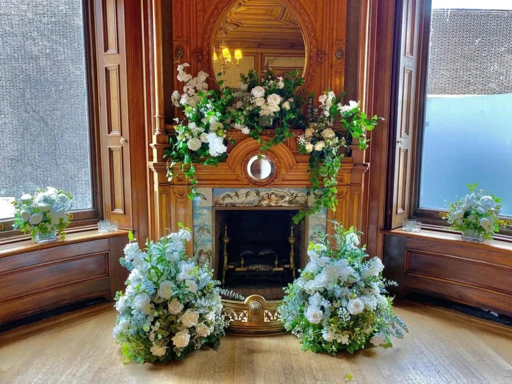The image features an elegant wedding decor setup by Casa D’Eramo in the wedding decor category. It shows an ornate wooden fireplace adorned with lush white and green floral arrangements, creating a classic and sophisticated atmosphere. The mantle is beautifully decorated with cascading flowers and foliage, complemented by tall windows allowing natural light to illuminate the display. Two large floral arrangements are symmetrically placed on the floor in front of the fireplace, enhancing the romantic and luxurious setting. This setup is ideal for a stylish and memorable wedding celebration.