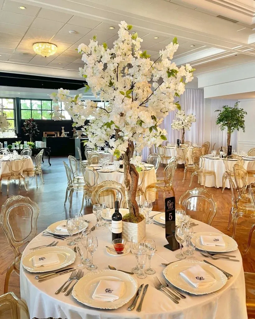 In this elegant wedding decor image from Casa D’Eramo, a beautifully set round table is featured in a sophisticated reception venue. The table is adorned with fine white linen and an exquisite tall floral centerpiece resembling a blossoming tree with white flowers. Place settings include neatly arranged plates, silver cutlery, and crystal glassware, each complemented by a menu card elegantly folded into the napkins. A bottle of wine stands next to a small bowl of sauce, adding to the inviting dining atmosphere. The chairs are stylishly transparent, adding a modern touch to the classic decor. The background reveals more tables similarly set up, contributing to the cohesive and luxurious ambiance perfect for a wedding celebration at Mariage Elegant, Canada.