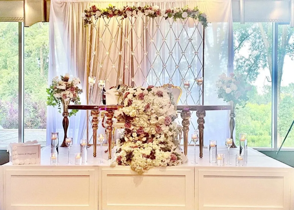 The image shows an elegant wedding decor setup by Casa D’Eramo, a vendor in the wedding category for Mariage Elegant, Canada. The backdrop features a luxurious arrangement of white and blush pink flowers cascading down a sophisticated table, accented by tall, ornate candlesticks. Delicate glass candle holders are placed symmetrically across the surface, creating a romantic ambiance. Behind the table is a lattice framework adorned with floral garlands, enhancing the elegant and serene setting. Large windows in the background offer a view of lush greenery, blending the outdoor beauty with the exquisite indoor decor.