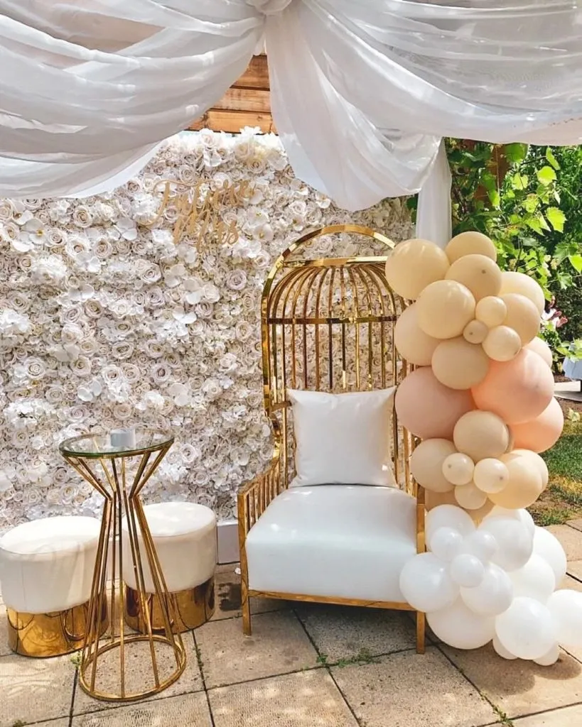 The image showcases a sophisticated wedding decor setup by Casa D’Eramo in the wedding decor category. A luxurious gold chair with a white cushion sits elegantly in front of a wall adorned with a lush display of white roses. To the right, a cascading arrangement of pastel-colored balloons, featuring shades of cream, peach, and white, adds a whimsical touch. Above, sheer white drapes create a soft and romantic canopy effect. A sleek gold and glass side table is positioned beside the chair, accompanied by two small white poufs, enhancing the setting's elegant ambiance, ideal for capturing memorable wedding moments.