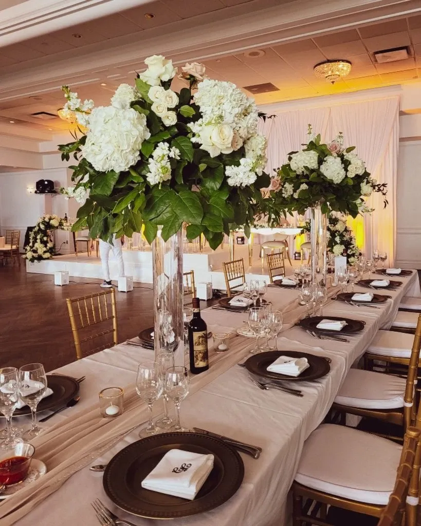 This image showcases a beautifully arranged wedding reception setup by Casa D’Eramo, featured in the Wedding Decor category of Mariage Elegant, Canada. Tall, elegant centerpieces consisting of lush white and cream flowers, including roses and hydrangeas, are placed in clear vases atop a long dining table. The table is adorned with white linens, gold-rimmed chargers, and neatly folded napkins, creating an elegant and sophisticated ambiance. Gold chiavari chairs complement the decor, and soft, ambient lighting enhances the romantic atmosphere of the venue. The backdrop features subtle drapery, adding to the luxurious feel of the setting.