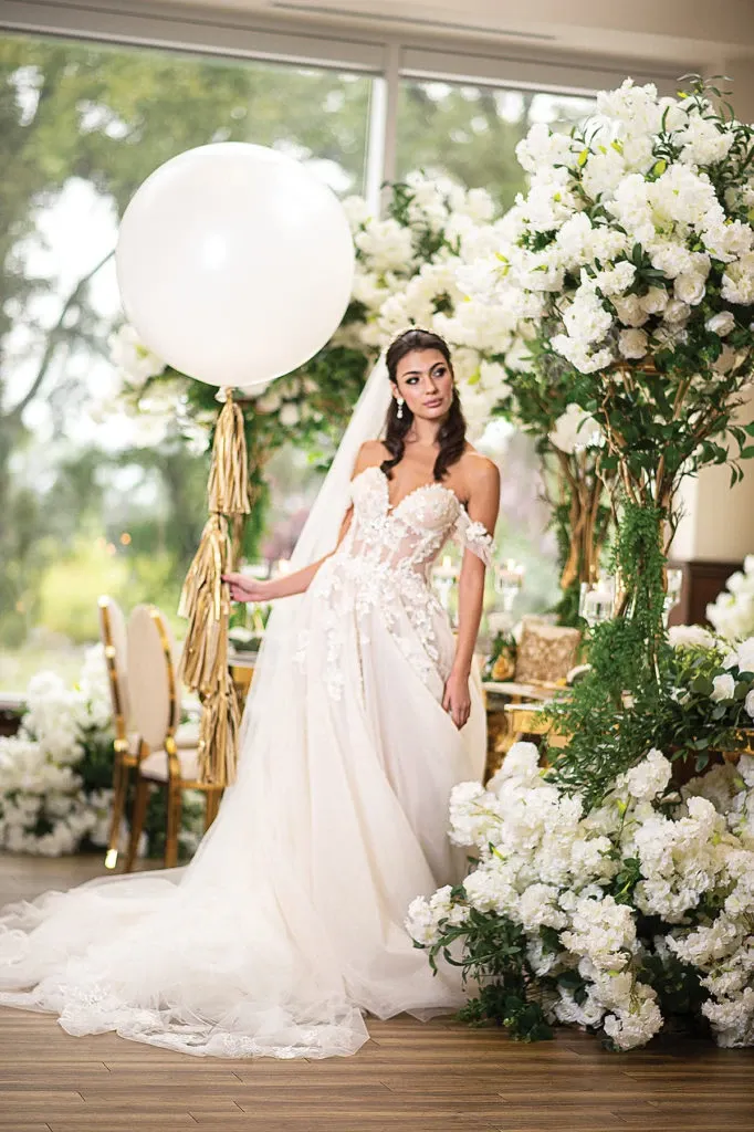 In this image from the wedding decor category by Casa D’Eramo, a bride stands elegantly in a beautifully arranged setting. She is wearing a detailed white gown with floral embellishments, complemented by a long, flowing veil. The decor includes lush arrangements of white flowers and greenery that frame the setting, creating a romantic and luxurious atmosphere. A large, white balloon with gold tassels adds a whimsical touch. The interior, with large windows, allows natural light to flood the space, enhancing the elegance of the decor. This setup is representative of a sophisticated wedding decoration theme.