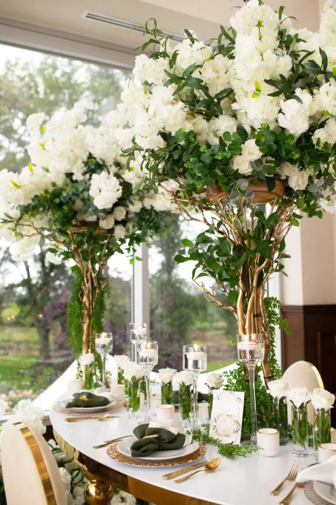 This image showcases an elegant wedding decor setup by Casa D'Eramo, featured in the wedding decor category at Mariage Elegant, Canada. The decor includes luxurious white flower arrangements with lush green foliage, artfully displayed on ornate gold stands. The table is adorned with white and gold tableware, complemented by green accents and delicate white candles, creating a sophisticated and romantic ambiance perfect for a wedding celebration.