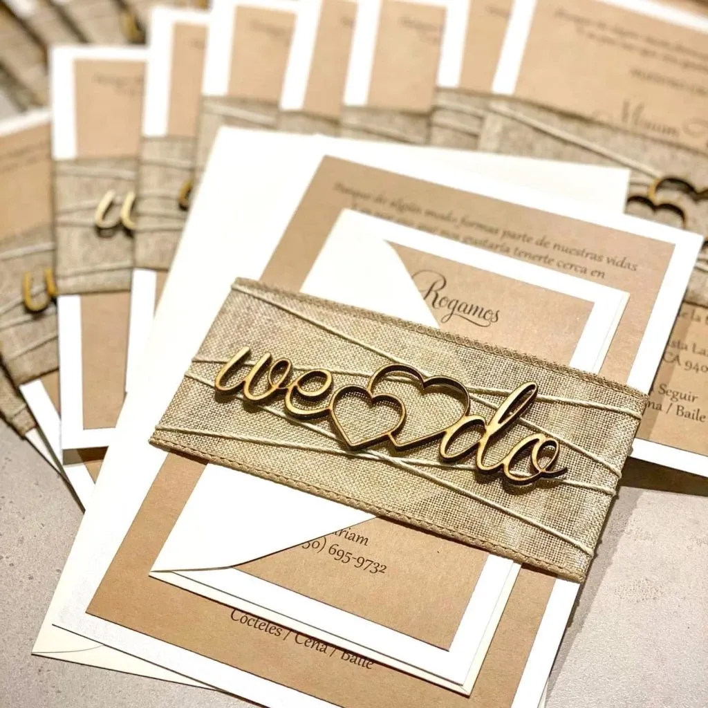 This image shows elegant wedding invitations designed by Casa D’Eramo, a vendor specializing in wedding decor for Mariage Elegant in Canada. The invitations feature a sophisticated earthy color palette with shades of beige and cream. Each invitation is meticulously crafted with a textured beige wrap around it, adorned with a gold-colored ornament that reads 'we ♥ do,' symbolizing love and union. The intricate details and refined style of the invitations highlight Casa D’Eramo's commitment to creating memorable and stylish pieces for wedding celebrations.