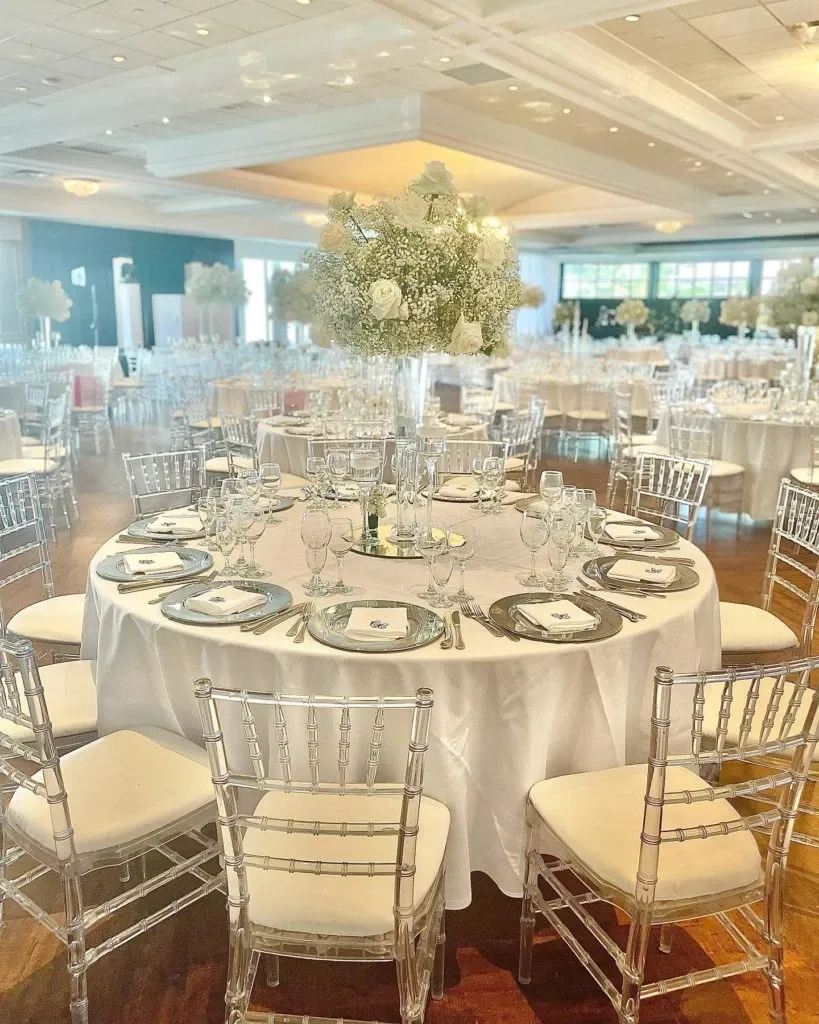 This image showcases an elegantly decorated wedding reception by Casa D’Eramo, featured in the Wedding Decor category on Mariage Elegant, Canada. The spacious venue is adorned with round tables draped in crisp white linens, each set for guests with fine china, crystal glassware, and neatly arranged silverware. The centerpiece of each table is a stunning floral arrangement featuring lush white roses and delicate baby's breath. The chairs are clear and modern, complementing the sophisticated ambiance. The soft lighting from the overhead fixtures casts a warm glow, enhancing the romantic and inviting atmosphere perfect for a memorable wedding celebration.