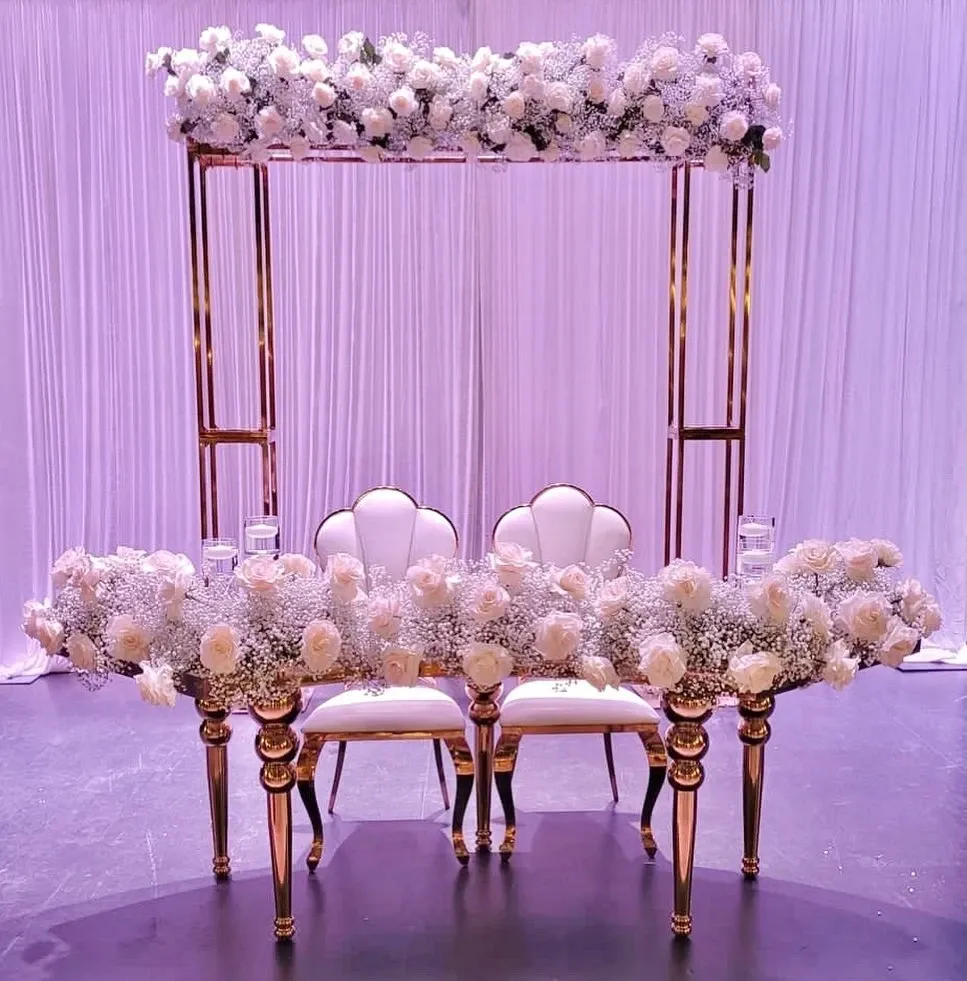 In this image, a luxurious wedding decor setup by Casa D’Eramo, a vendor under the Wedding Decor category, is showcased. The arrangement features an elegant sweetheart table adorned with lush white roses and delicate baby's breath flowers, creating a romantic atmosphere. The table is complemented by two sophisticated, white upholstered chairs with gold accents. Behind the table, a stylish backdrop of cascading white fabric adds depth and enhances the overall aesthetic, while a golden metallic structure with additional floral arrangements crowns the setting. This elegant design exemplifies the high-quality wedding decor services offered by Casa D’Eramo for Canadian weddings.