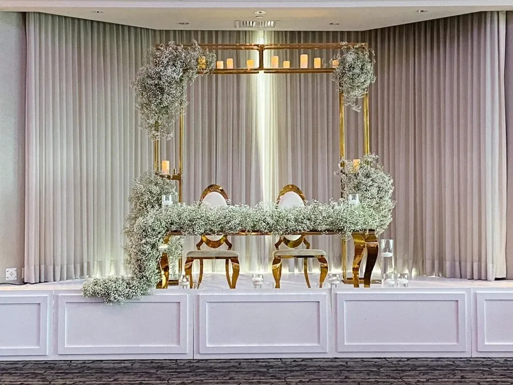 The image showcases an elegantly arranged wedding decor setup by Casa D’Eramo, featured in the wedding decor category on Mariage Elegant, Canada. The focal point is a beautifully adorned sweetheart table with two ornate chairs, framed by a luxurious backdrop of floor-to-ceiling curtains. A stunning canopy structure, decorated with delicate white florals and cascading greenery, highlights the romantic atmosphere. Soft candlelight adds a warm glow, enhancing the sophisticated and intimate ambiance ideal for a dream wedding celebration.