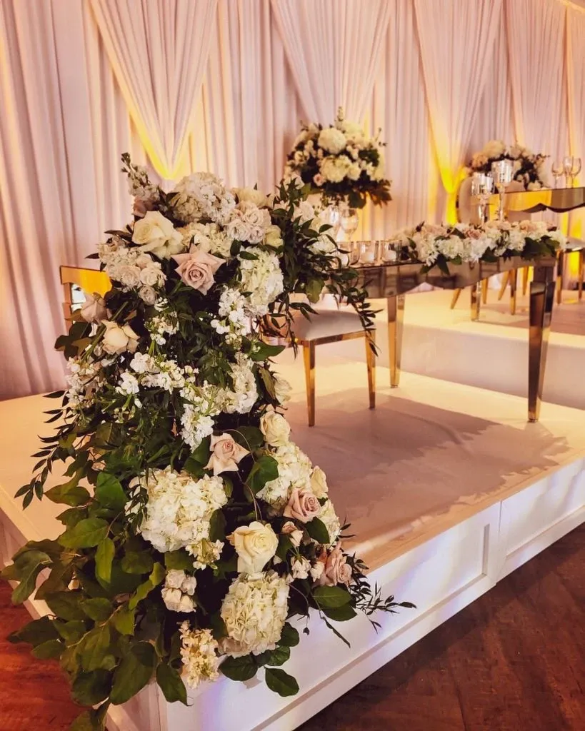 The image features a luxurious wedding decor setup by Casa D'Eramo, categorized under wedding decor. It showcases an elegant arrangement with cascading floral decorations, prominently displaying white and blush roses, hydrangeas, and lush greenery elegantly draped on a raised platform. Golden accents highlight the table and chairs placed on the platform, adding a touch of sophistication. White draped fabric serves as a backdrop, with soft, warm lighting creating a romantic ambiance. This decor exemplifies the refined and elegant style that Casa D'Eramo offers for weddings in Canada.