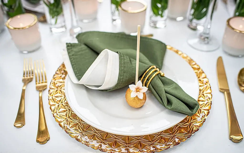 This image showcases an elegantly arranged wedding table setting presented by Casa D’Eramo, a vendor in the Wedding Decor category for Mariage Elegant in Canada. The centerpiece features a white plate with an intricately designed gold charger beneath, complemented by gold cutlery. On the plate, a two-toned napkin—dark green and white—is artistically folded and secured with gold napkin rings. A decorative element, resembling a small golden ornament with a single delicate white flower, is placed atop the napkin. Surrounding the centerpiece are multiple small candles in glass holders, adding a warm, romantic ambiance perfect for a sophisticated wedding celebration.