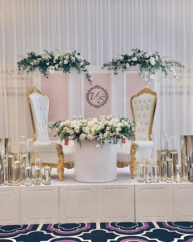 The image showcases an elegant wedding decor setup by Casa D’Eramo, a vendor in the wedding decor category. The scene features two luxurious, white upholstered chairs with ornate gold detailing, positioned on a raised platform. Between the chairs, a large arrangement of white flowers is beautifully displayed in a cylindrical vase. Surrounding the platform are multiple glass pillars of varying heights, each holding a tall candle, adding a touch of sophistication. Above the chairs are two lush floral arrangements with white and green foliage, enhancing the romantic atmosphere. In the center background, a decorative monogram featuring stylized initials adds a personalized touch to the elegant setting. This decor setup creates a regal and inviting ambiance for a wedding celebration.