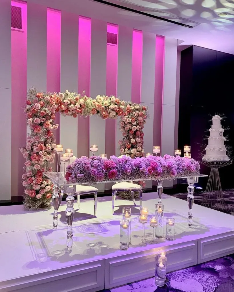 The image showcases an elegant wedding decor setup by Casa D’Eramo, categorized under Wedding Decor for Mariage Elegant, Canada. The focal point is a beautifully crafted floral arch composed of pink, white, and blush roses, providing a stunning backdrop. In front of the arch, there are two sophisticated white chairs paired with a table adorned with similar floral arrangements. The ambient lighting casts a soft, romantic glow with a purple hue, enhancing the dreamy atmosphere. To the right, a tall, intricately designed white wedding cake is displayed on a pedestal, complementing the overall decor. Crystal candlesticks with glowing candles are artfully placed around the setting, adding an additional layer of elegance and warmth to the composition.