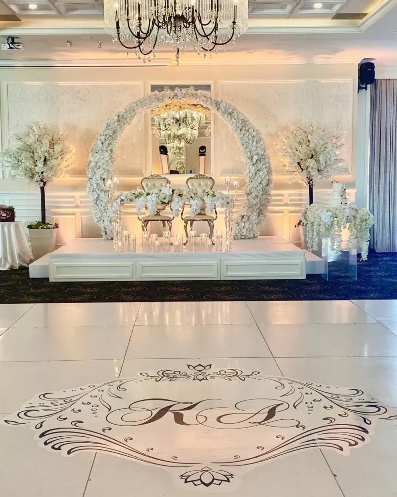 This image showcases an elegant wedding decor setup by Casa D’Eramo in Canada. The centerpiece features a luxurious circular floral arch adorned with white roses, beautifully framing two ornate chairs on a white platform. Tall floral arrangements complement the scene, alongside cascading candle displays adding a romantic ambiance. The floor displays monogrammed initials 'K' and 'P,' suggesting a personalized touch for the bride and groom. A stunning chandelier and soft drapery complete the sophisticated atmosphere, capturing the essence of exquisite wedding decoration.