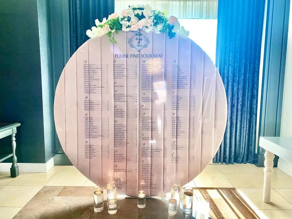 This image features a beautifully arranged wedding seating chart designed by Casa D’Eramo in the wedding decor category. The seating chart is crafted on a large, elegant round board with a subtle off-white color, providing a soft backdrop for the neatly organized lists of guest names. At the top, delicate floral arrangements with pink and white flowers add a romantic touch. Below, several glass votive candles are placed on the floor, creating a warm, inviting ambiance. This exquisite setup is ideal for guiding guests to their seats at a wedding, showcasing Casa D’Eramo's expertise in wedding decor.