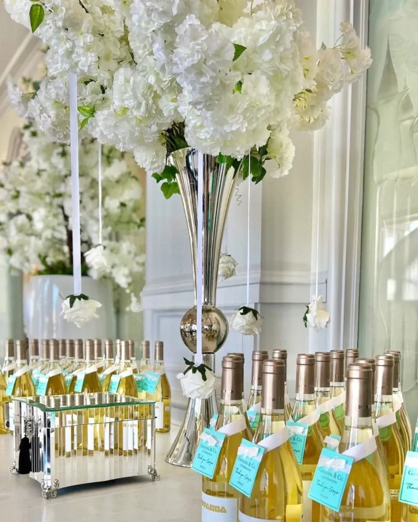 The image showcases an elegant wedding decor setup by Casa D’Eramo, a vendor listed in the Wedding Decor category on Mariage Elegant, Canada. Prominently featured is a large, sophisticated silver vase filled with lush white flowers, exuding a sense of luxury and romance. Below the vase, numerous bottles of white wine are neatly arranged, each adorned with a blue tag, possibly indicating personalized messages or guest seating arrangements. The setup is completed with smaller flower arrangements, creating a cohesive and stylish display perfect for a refined wedding celebration.
