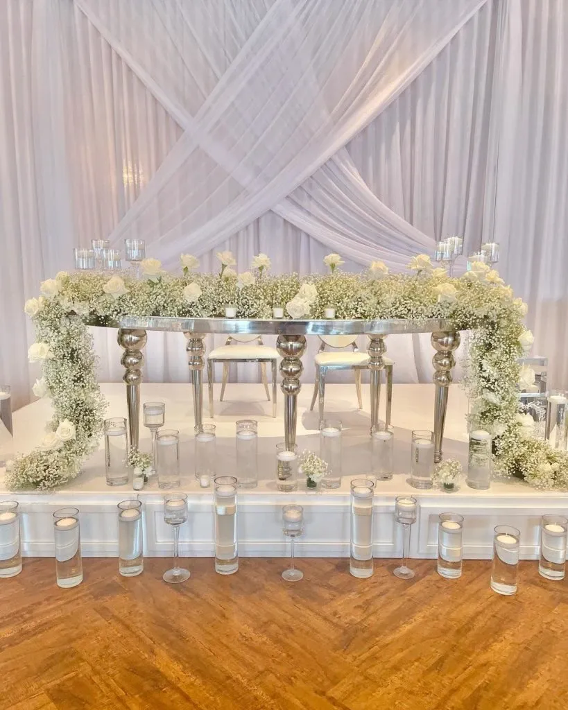 This image showcases a beautifully arranged wedding decor setup by Casa D’Eramo, featured in the wedding decor category at Mariage Elegant, Canada. The scene presents an elegant, elevated table adorned with an abundance of white roses and delicate baby's breath, creating a lush floral display. The backdrop features softly draped white fabric, adding a sophisticated and serene ambiance. Cylindrical glass vases filled with water and floating candles are artistically arranged around the base, enhancing the romantic and tranquil atmosphere perfect for a wedding celebration.