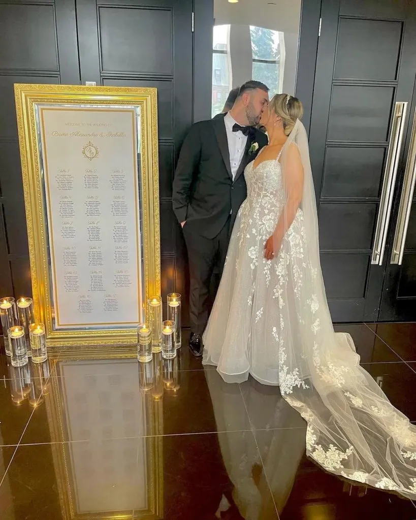 In this image, a newlywed couple shares a kiss in front of an elegant wedding seating chart, crafted by Casa D’Eramo, a vendor specializing in wedding decor. The bride is dressed in a flowing white gown with intricate lace detailing, accompanied by a long veil, while the groom wears a classic black tuxedo. The seating chart is framed in an ornate gold frame and is adorned with a sophisticated design, listing the names of guests in an organized manner. Surrounding the chart, an array of candles in glass holders adds a romantic ambiance, reflecting light onto the polished dark floor. This setting showcases Casa D’Eramo’s expertise in creating luxurious and elegant wedding decor.