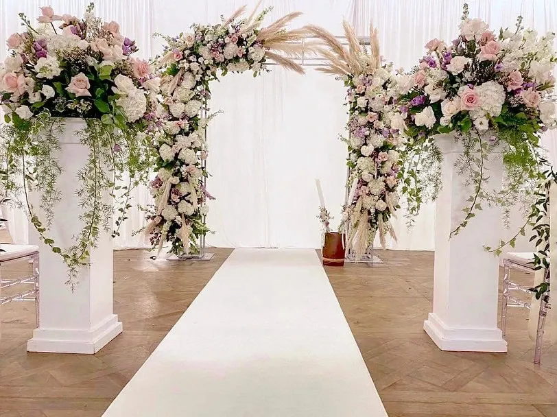The image showcases an elegant wedding decor setup by Casa D’Eramo, featured in the Wedding Decor category on Mariage Elegant, Canada. The focal point is a beautifully decorated ceremonial arch adorned with lush flowers in soft pastel hues, including roses and hydrangeas. Flanking the arch are tall white pedestals, each topped with cascading floral arrangements and greenery, creating a romantic and enchanting atmosphere. A white aisle runner completes the setup, leading to the arch, perfect for a sophisticated wedding ceremony.