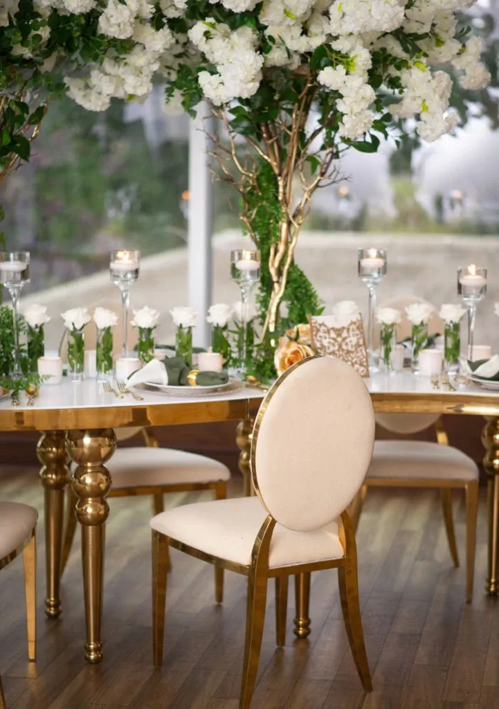 The image showcases an elegant wedding decor setup by Casa D’Eramo, categorized under Wedding Decor, featured on Mariage Elegant, Canada. The scene displays a beautifully arranged table with golden legs and a glossy finish, adorned with fresh white roses and lush greenery. Tall glass candle holders with lit candles provide a warm ambiance, while luxurious gold-trimmed chairs complement the sophisticated setting. A backdrop of large floral arrangements with white blooms and green leaves adds an opulent touch to the romantic wedding atmosphere.