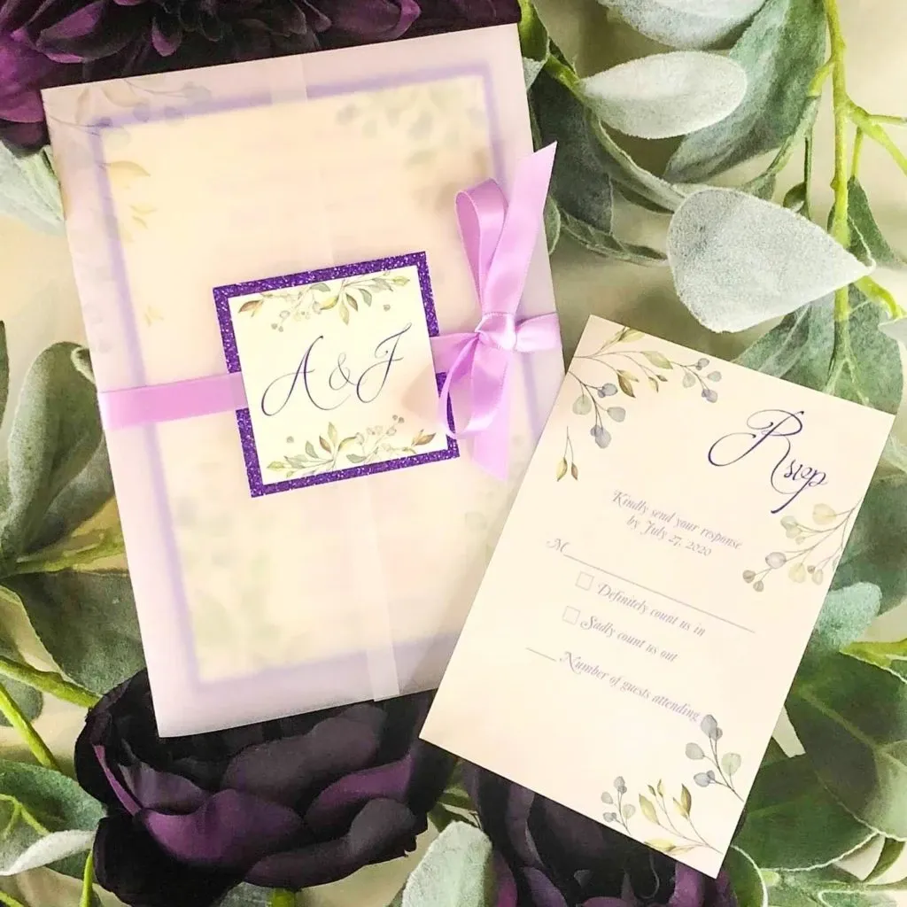 The image features elegant wedding invitations designed by Casa D’Eramo in the Wedding Decor category for Mariage Elegant, Canada. The invitation set includes a beautifully crafted card adorned with lavender and green floral accents, wrapped with a sheer purple ribbon and a sparkly square label with the initials “A & J.” Beside it lies a matching RSVP card, requesting guests to respond by a specified date. The items are artfully arranged on a backdrop of lush purple and green foliage, creating a sophisticated and inviting presentation ideal for any wedding occasion.