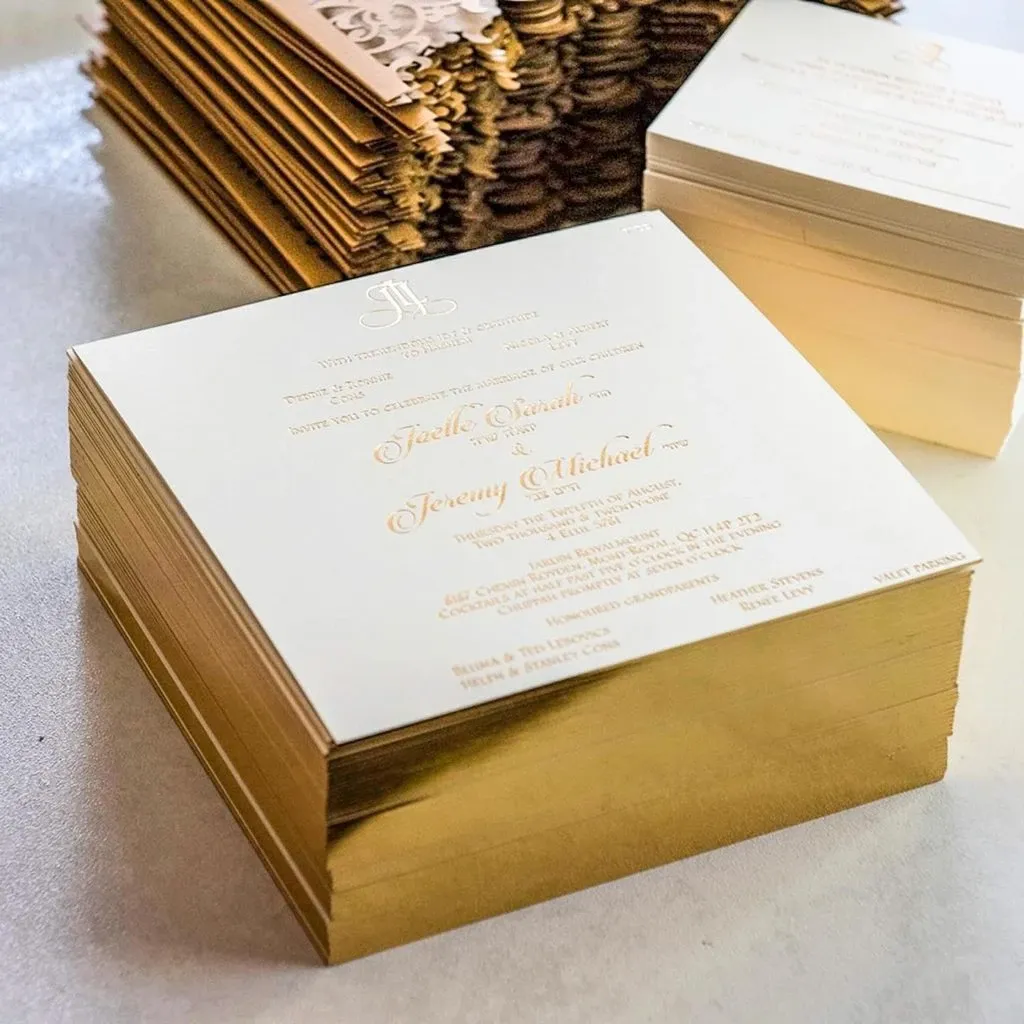 The image shows a stack of elegantly designed wedding invitations by Casa D’Eramo, categorized under Wedding Decor. The invitations feature a sophisticated gold foil border and intricate lettering, highlighting the couple's names, Jocelyn Emma and Jeremy Michael, with a detailed decorative header. The luxurious invitations are set on a white table, exemplifying Casa D’Eramo's expertise in creating high-quality, bespoke wedding stationery, perfect for stylish and memorable nuptials.