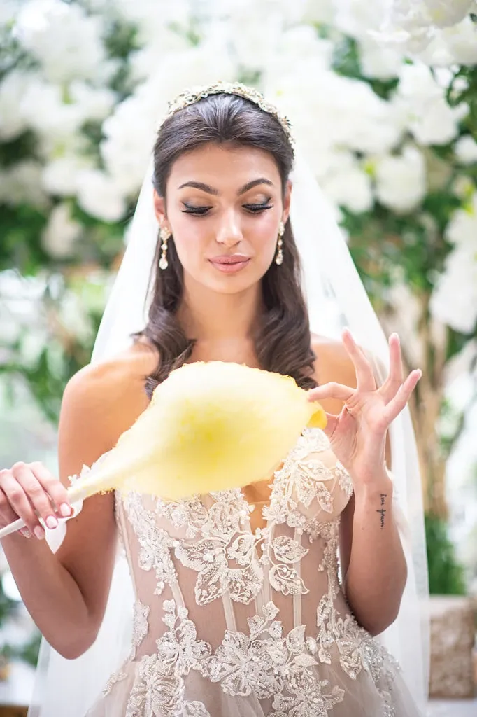 A bride elegantly dressed in a lace wedding gown holds a fluffy piece of yellow cotton candy. She stands amidst lush greenery and white floral arrangements, enhancing the romantic atmosphere. Her hair is styled with a delicate tiara and she wears drop earrings. This image showcases the exquisite decor by Casa D’Eramo, a vendor specializing in wedding decor, at Mariage Elegant, Canada.