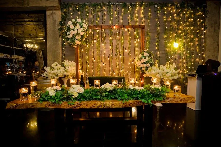 A beautifully arranged wedding decor setting by Casa D’Eramo, showcasing their expertise in the Wedding Decor category. The image features an elegantly adorned table with a rich wooden surface covered in lush greenery and interspersed with numerous white floral arrangements. Surrounding the centerpiece are a variety of warm, glowing candles that add a romantic ambiance to the setting. Behind the table, twinkling string lights drape the wall, creating a magical and inviting backdrop. The overall theme exudes elegance and sophistication, perfectly aligning with wedding celebrations in Canada.