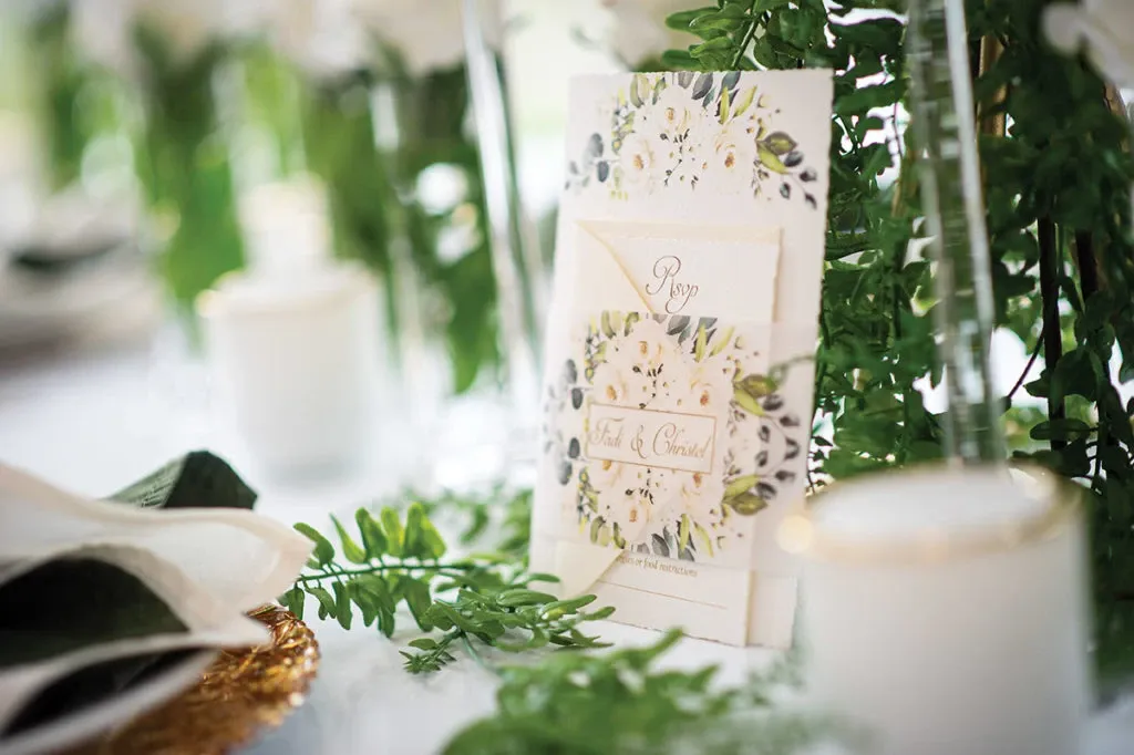 This image showcases an elegant wedding decor setup by Casa D’Eramo in the wedding decor category. The focus is on a beautifully designed RSVP card featuring floral patterns, set amidst lush green foliage, creating a fresh and sophisticated look. A soft white candle adds to the ambiance, while a gold-rimmed glass and a delicately folded napkin contribute to the table's refined aesthetic. The elements reflect Casa D’Eramo’s attention to detail and expertise in creating stylish and inviting wedding settings, perfect for a memorable celebration in Canada.