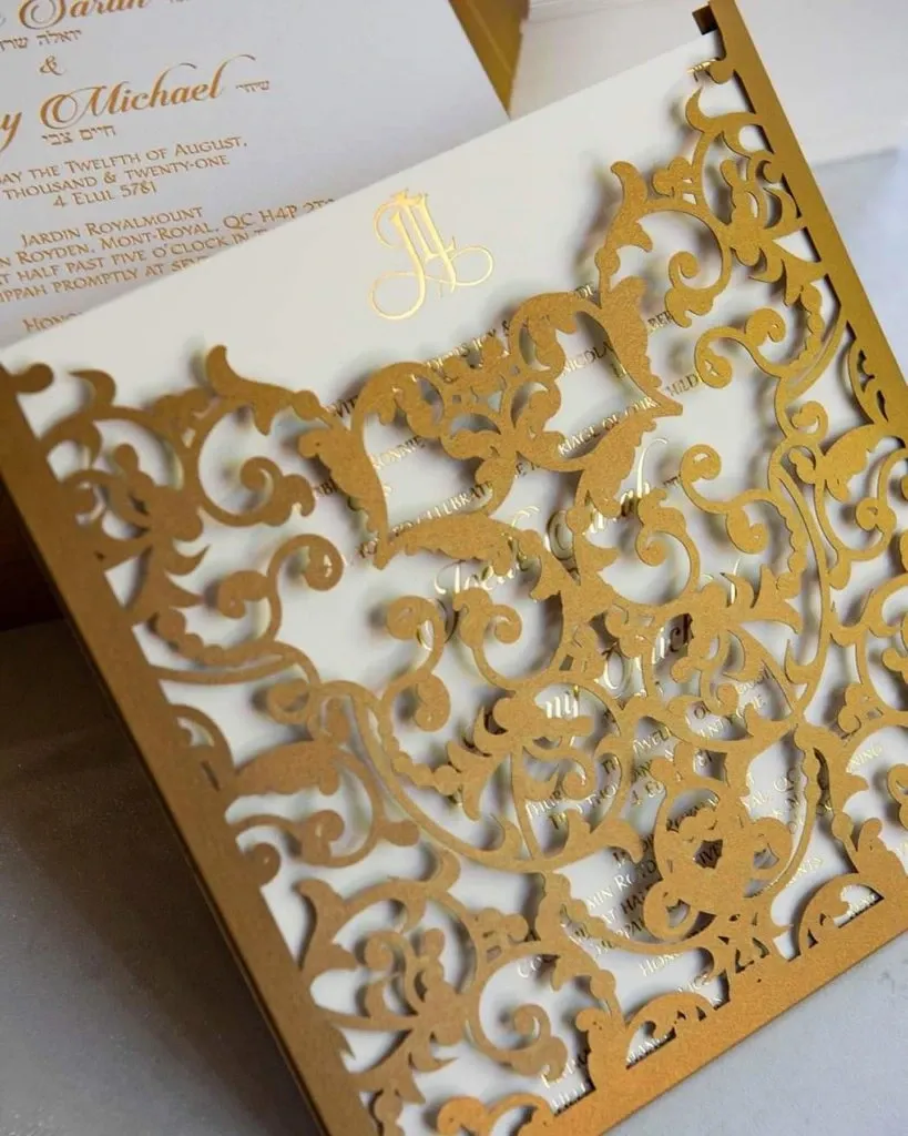 The image showcases an intricately designed wedding invitation by Casa D’Eramo, categorized under Wedding Decor. The centerpiece is an elegant gold laser-cut overlay featuring ornate floral and scroll motifs, providing a glimpse of the classic text and elegant font of the invitation underneath. The exquisite craftsmanship adds a touch of sophistication and luxury, ideal for any elegant wedding. The gold and white color palette is both timeless and versatile, complementing various wedding themes. This invitation is a prime example of the high-quality wedding decor options offered by Casa D’Eramo.